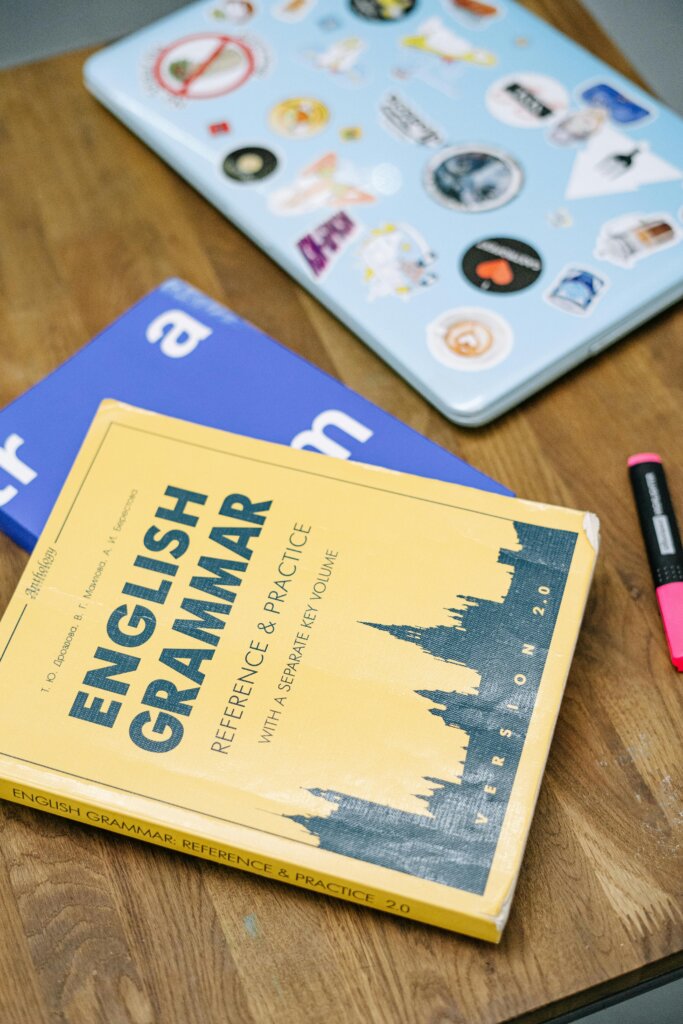 pexels-photo-5676740-5676740 High-angle view of an English grammar book and a laptop with stickers on a wooden desk.